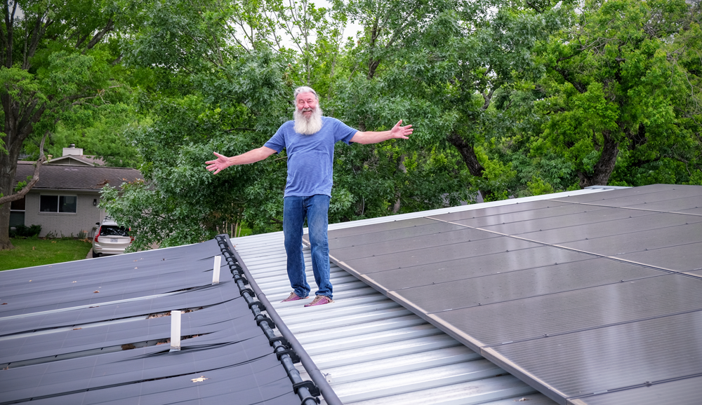 Jerry with his arms outstretched standing on top of a roof. There is a solar heating system installed on his left and a solar energy system on his right.