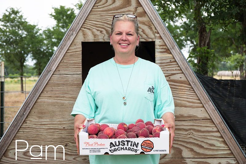 Pam holding a box of peaches standing in front of a small triangular shed or chicken coop. 