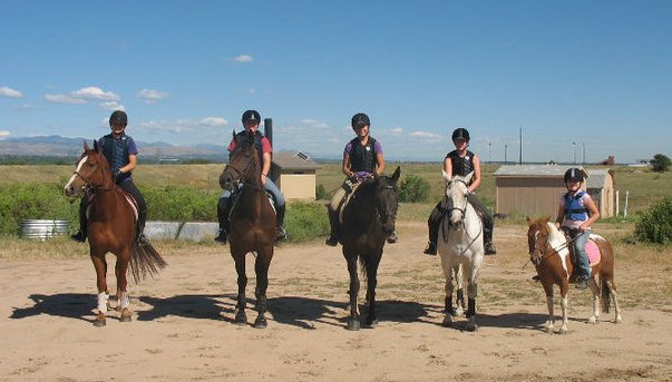 Six people on horses, ont he far right, a child is riding a pony. There is blue sky and mountains in the background.